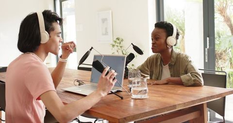 Two co-hosts recording conversational podcast in bright home studio with laptop and mics