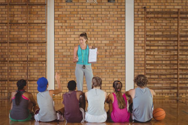 Female coach mentoring high school students in a basketball court. Coach holding a clipboard and explaining strategies to the students sitting on the floor. Ideal for use in educational materials, sports training guides, teamwork and leadership articles, and youth sports promotion.