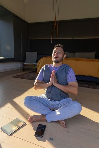 Asian man meditating on sunlit bedroom floor with smartphone and notebook, mindfulness