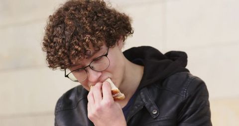 Curly-haired young man wearing glasses leather jacket biting sandwich outdoors