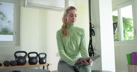 Woman using tablet in bright home gym wearing green top and grey leggings