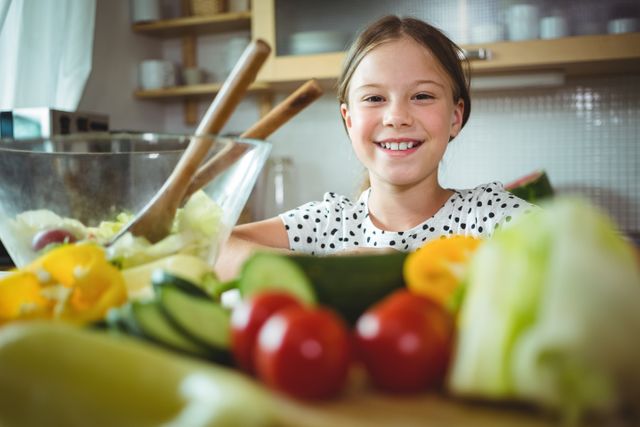 Young girl smiling while preparing a fresh salad in a home kitchen. Ideal for use in content related to healthy eating, family activities, children's nutrition, and home cooking.