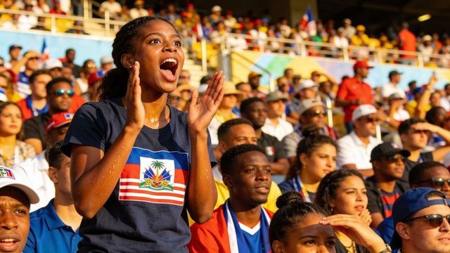 Energetic haitian teen cheering in sunlit stadium crowd wearing national emblem tee