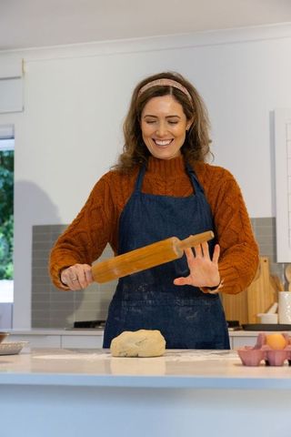 Smiling woman rolling dough on kitchen island wearing denim apron and cozy sweater
