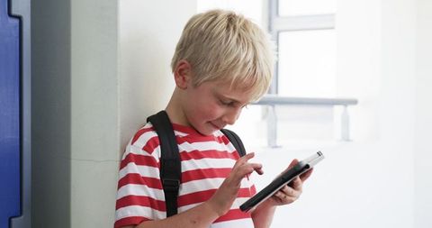 Blond schoolboy using tablet while leaning in bright corridor with backpack