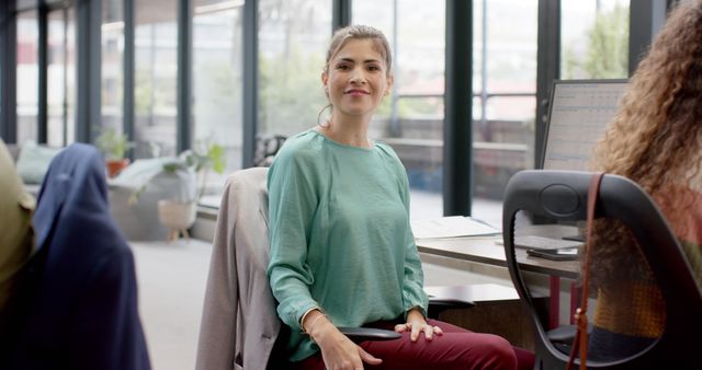 Businesswoman in a modern office environment sitting at her desk, smiling confidently at the camera. She wears casual attire, showcasing a relaxed yet professional vibe. Ideal for depicting workplace culture, productivity, and professional success in corporate or creative fields. Suitable for use in business website imagery, career-oriented promotions, and workplace inclusion materials.