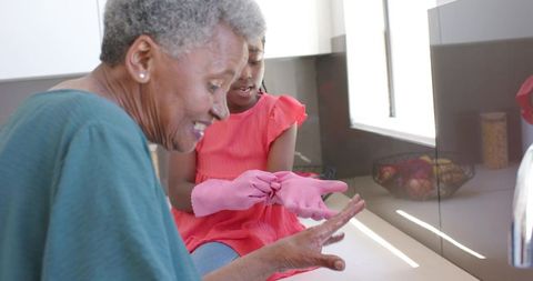 African american grandmother helping granddaughter clean kitchen wearing pink gloves