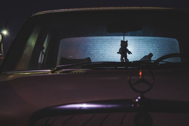 Interior view of a vintage car at night with a plush toy hanging from the rearview mirror. Ideal for use in themes of nostalgia, retro vibes, or vintage transportation. Can also be used in mood-setting visuals, nighttime scenes, or automotive-related content.