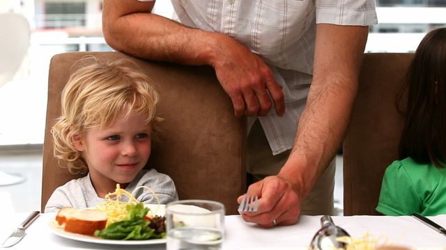 Father bending over to persuade child to eat vegetables during lunch at home. Ideal for use in articles about parenting, healthy eating, family meals, encouraging children to try new foods, or portrayals of family life.