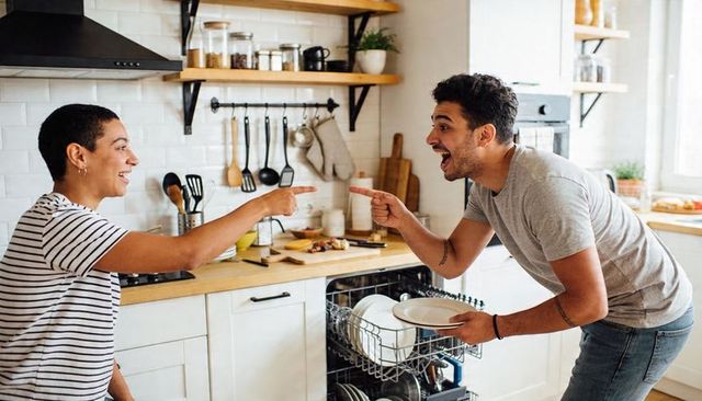Playful couple laughing and pointing while loading dishwasher in bright modern kitchen