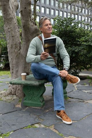 Middle-aged man reading on green stone bench with coffee in urban courtyard
