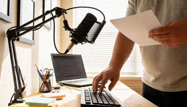 Man recording podcast at home desk with microphone and laptop backlit by blinds