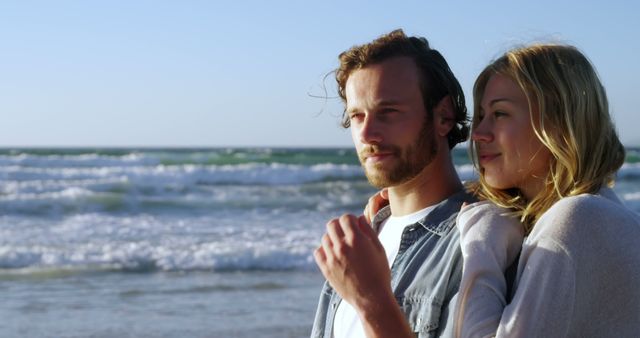 Young couple standing close to each other, gazing at the ocean waves and enjoying the sunset. The serene setting and the romantic moment make it ideal for travel and tourism content, relationship and love themes, or promotional material for beach getaways.