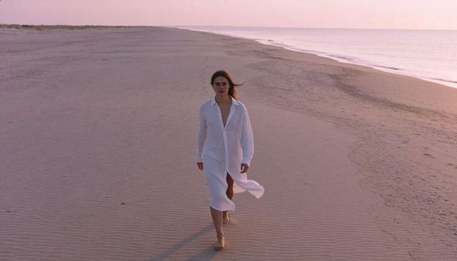Solitary woman walking on rippled sand wearing white shirt dress at pastel sunrise beach