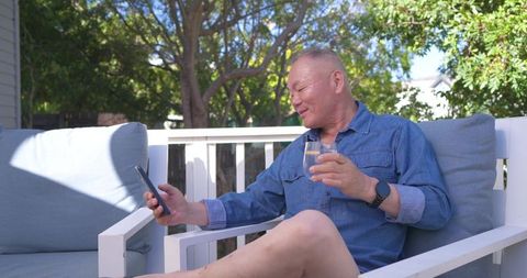 Relaxing mature asian man scrolling smartphone on sunny backyard deck holding glass of water