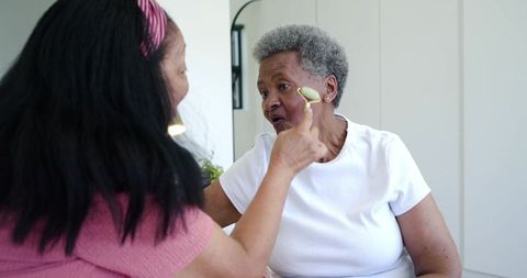 African american caregiver applying facial roller on senior woman during skincare routine
