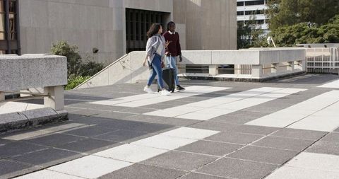 Young diverse professionals walking on urban rooftop plaza pulling suitcase and using phone