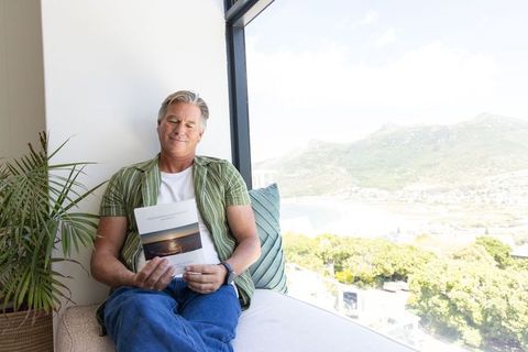 Senior man relaxing on window seat reading booklet with serene coastal bay view