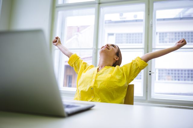 Young businesswoman in a yellow shirt stretching with outstretched arms at her desk in an office. She looks excited and happy, possibly celebrating a success or taking a break. Ideal for use in business, motivation, and workplace wellness contexts.