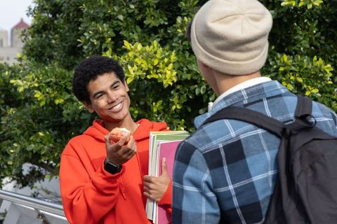Smiling teen in orange hoodie offering apple while chatting with friend on sunlit campus