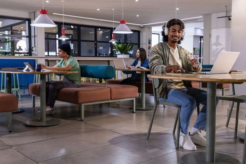 Indian man wearing headphones working on laptop in coworking cafe with diverse freelancers