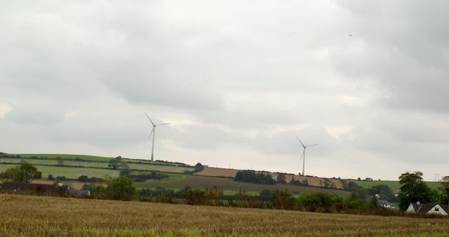 Wind turbines on rolling green hills under cloudy sky. Suitable for themes of renewable energy, environmental conservation, rural scenery, and agriculture. Ideal for illustrating countryside landscapes, climate action initiatives, and sustainable energy projects.