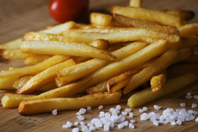 Crispy golden French fries seasoned with sea salt, served on a wooden table with a cherry tomato in the background. Ideal for illustrating food blogs, restaurant menus, snack advertisements, or social media posts about delicious comfort food and fast-food delights.