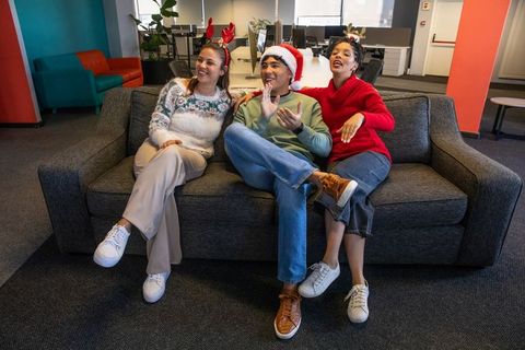 Diverse coworkers celebrating casual office holiday on sofa wearing Santa hat and reindeer antlers