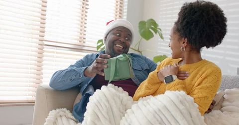 African american couple exchanging green gift on sofa wearing santa hat, cozy holiday joy