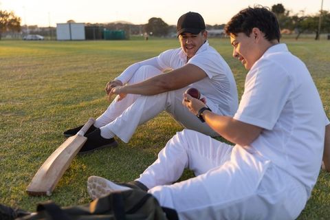 Cricket teammates sitting on grass at golden hour inspecting ball and bat, pregame chat