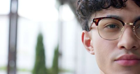 Young man wearing tortoiseshell glasses gazing forward in bright corridor with greenery