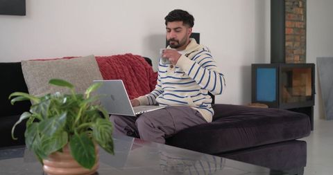 Indian man sipping coffee while working on laptop in modern minimalist living room