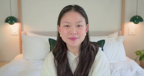 Asian woman sitting on bed in cozy modern bedroom wearing hoodie and gold earrings