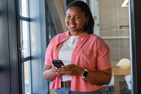 African american professional woman smiling holding flip phone by window in modern office