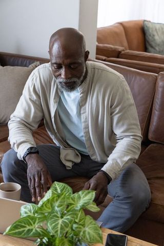 Senior african man sitting on leather sofa using laptop in cozy sunlit living room