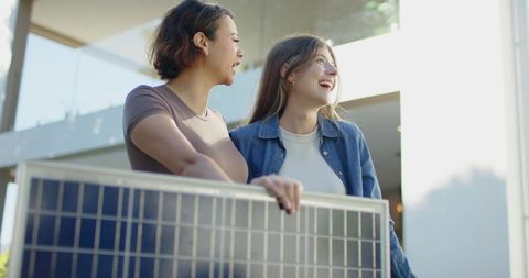 Young women holding solar panel on modern balcony smiling and planning solar installation