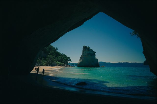Tourists exploring picturesque beach and ocean, viewed from inside cave, creating dramatic light contrast. Ideal for travel blogs, adventure advertising, magazine covers highlighting scenic destinations.