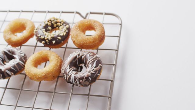 Assorted donuts with various toppings cooling on wire rack create a visually appealing scene against a white background. Ideal for illustrating bakery promotional materials, food blogs, recipe books, and dessert menus. Perfect for conveying themes of indulgence, gourmet treats, and homemade baking.