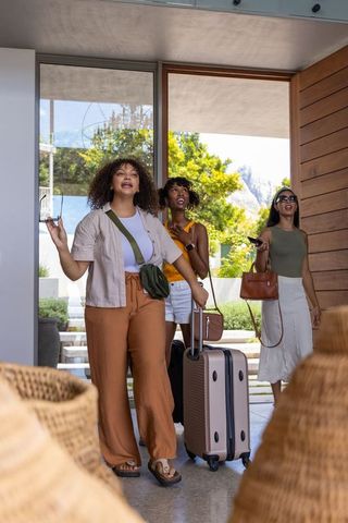 Multiracial friends arriving with rolling luggage at stylish modern vacation home entrance