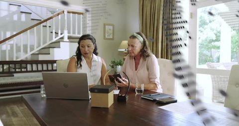 Two women collaborating at home dining table using laptop and charging smartphone