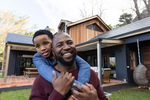 African american father carrying son on back outside modern wood house smiling together