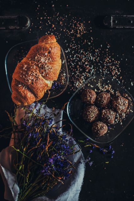 High-angle view of rustic breakfast foods including a croissant and chocolate treats on dark wooden table. Bouquets of flowers add aesthetic touch. Ideal for use in bakery promotion, breakfast menus, or food blog content.