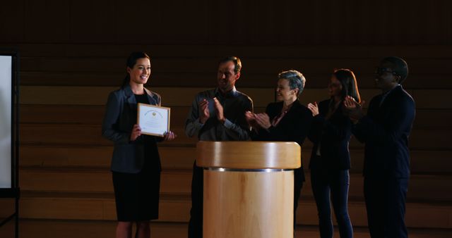 Woman in a formal suit receiving a business award from a colleague on stage, while others applaud. Suitable for presentations, business success stories, employee recognition programs, teamwork promotions, and corporate event highlights.