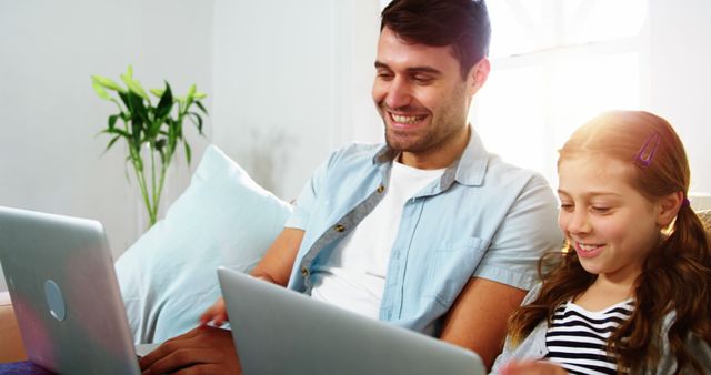 Image showing father and daughter sitting on a couch using laptops. Ideal for use in contexts such as family bonding, home learning, technology use in family settings, and promoting parent-child fun activities.