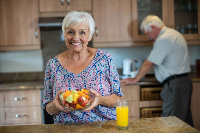 Senior woman smiling and holding a bowl of fresh fruit in a modern kitchen, with a man working in the background. Ideal for use in advertisements promoting healthy eating, active lifestyles for seniors, retirement living, and domestic life. Perfect for illustrating concepts of happiness, togetherness, and wellness in older adults.