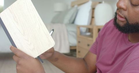 African american man sitting on bed holding wooden journal and pen, writing in bedroom