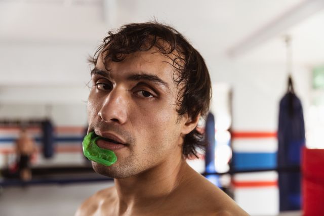 Portrait close up of a young biracial male boxer with short dark hair, shirtless with a gumshieldin his mouth, standing in a boxing gym looking straight to the camera.