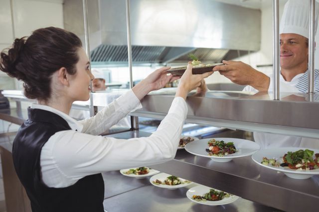 Chef handing a prepared dish to a waitress at the order station in a commercial kitchen. Ideal for use in articles or advertisements related to restaurant operations, culinary arts, teamwork in hospitality, and professional kitchen environments.