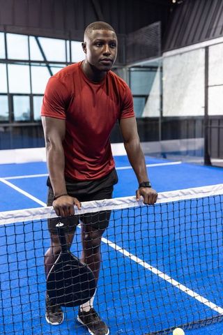 African american athlete leaning on paddle tennis net holding racket on blue indoor padel court