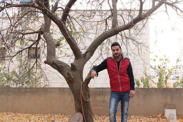 Young man wearing red vest and jeans standing casually near a tree shedding leaves in an urban setting. Ideal for themes like autumn fashion, nature, casual lifestyle, or urban parks.
